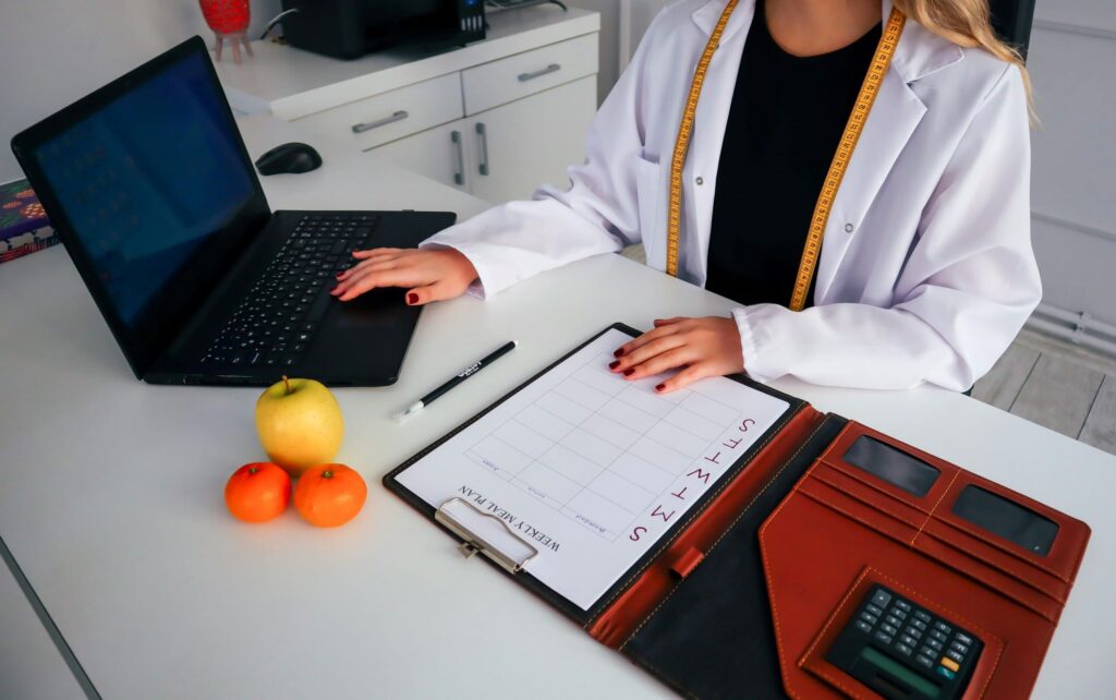 La dietista è alla scrivania davanti al pc ad elaborare la dietoterapia. Sulla scrivania ci sono calcolatrice, dieta e dei mandarini.
